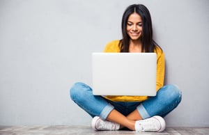 Happy young woman sitting on the floor with crossed legs and using laptop on gray background1